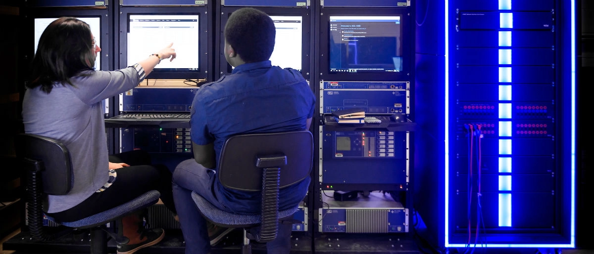Two people sitting down in front of many screens