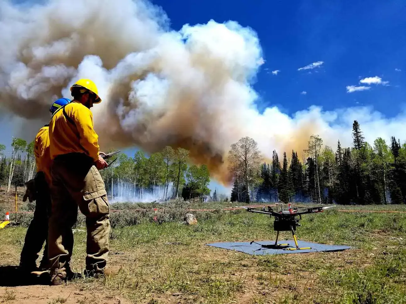 Two men stand before a drone with smoke in the background. 