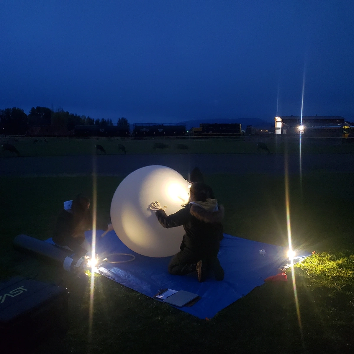 People blow up a white weather balloon at night.