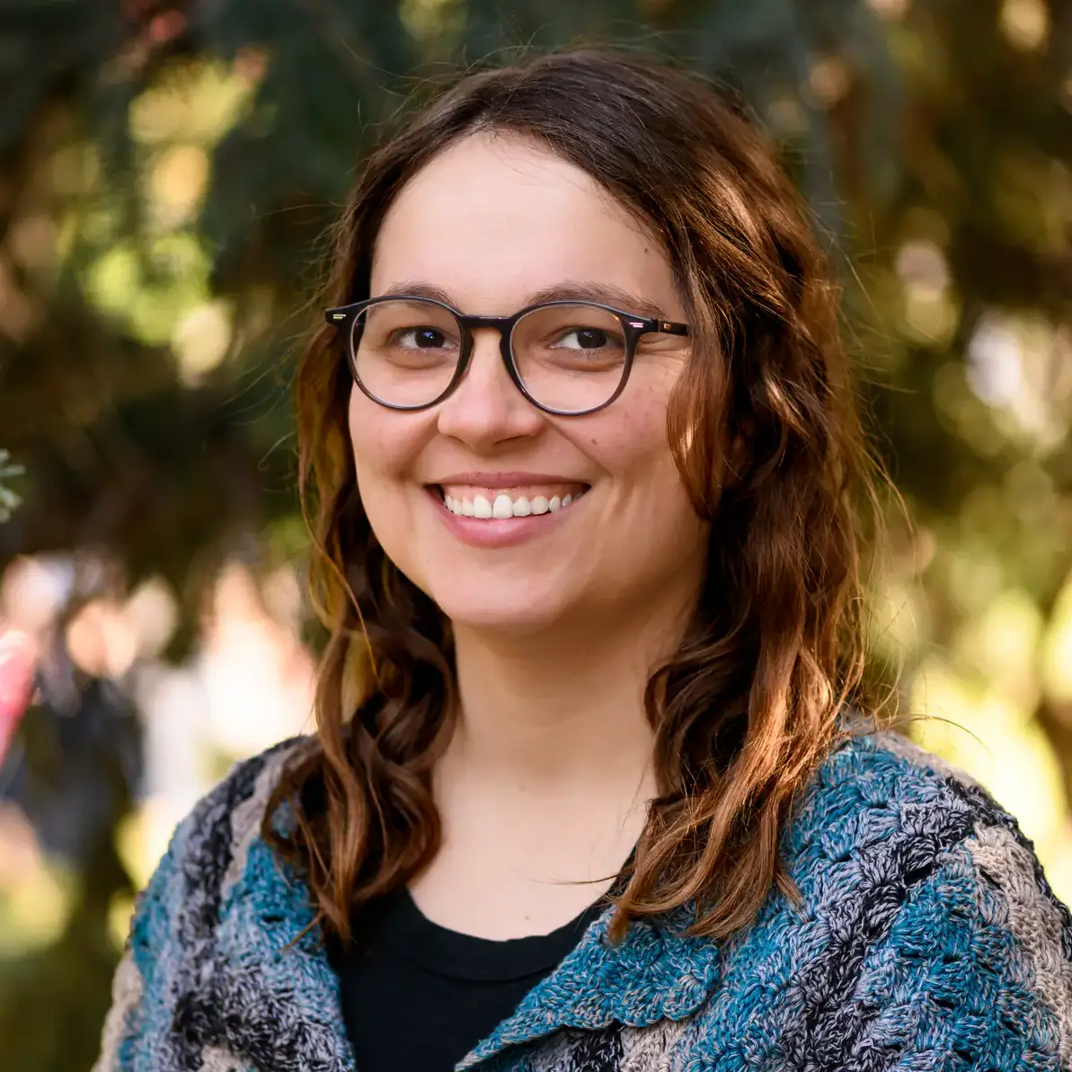 A person with glasses smiles outdoors with trees in the background.