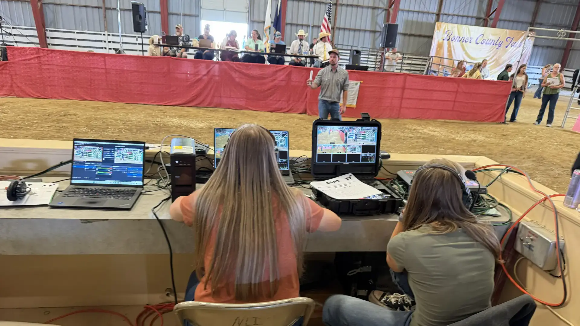 Two girls sit at a table with computers adjacent to a dirt arena.