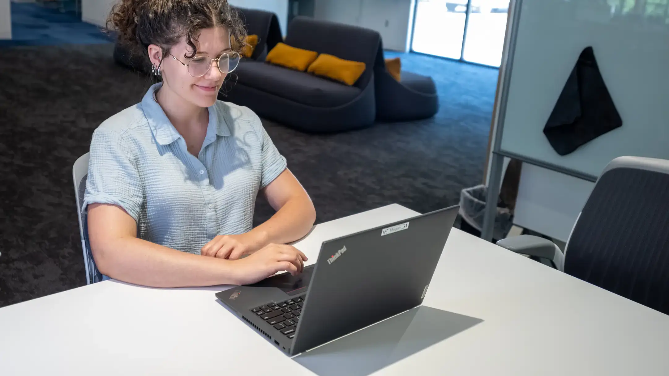 A person sits at a desk using a laptop in a modern workspace with chairs and whiteboards in the background.
