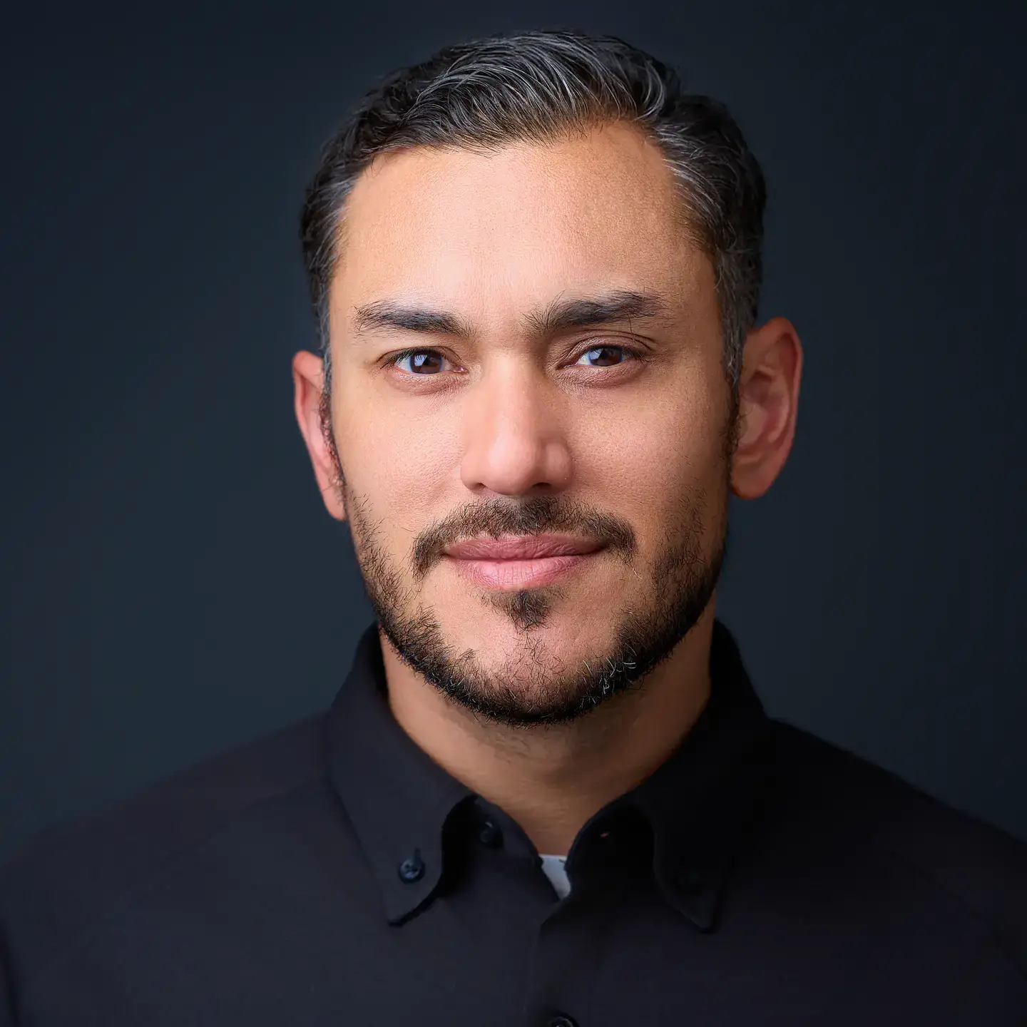 Professional headshot of a man with short dark hair and a trimmed beard, wearing a black collared shirt.