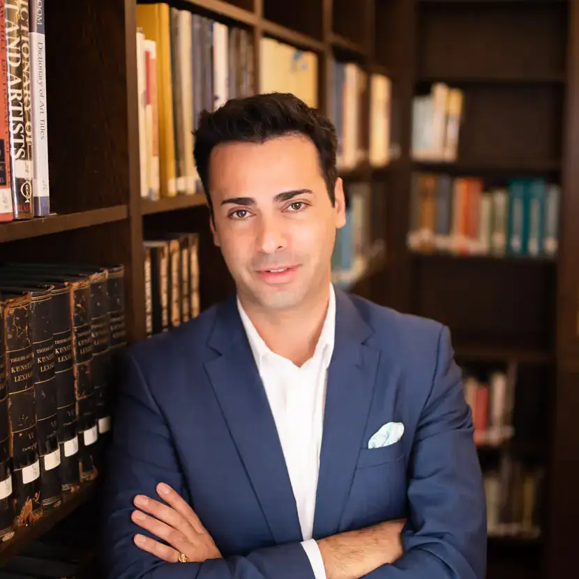 A person in a blue suit stands with arms crossed in front of a bookshelf in a library.