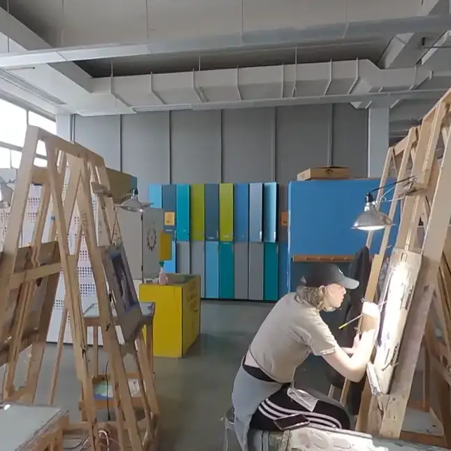 Person with a paintbrush leaning into an easel working on an art project, in a room with other easels and colorful lockers.