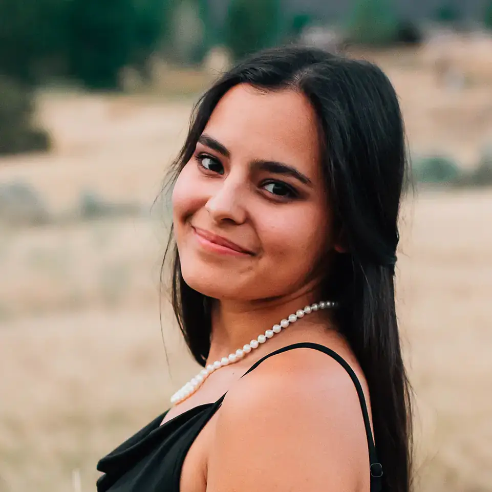 Young woman in a black dress and pearl necklace smiles outdoors in a grassy field.