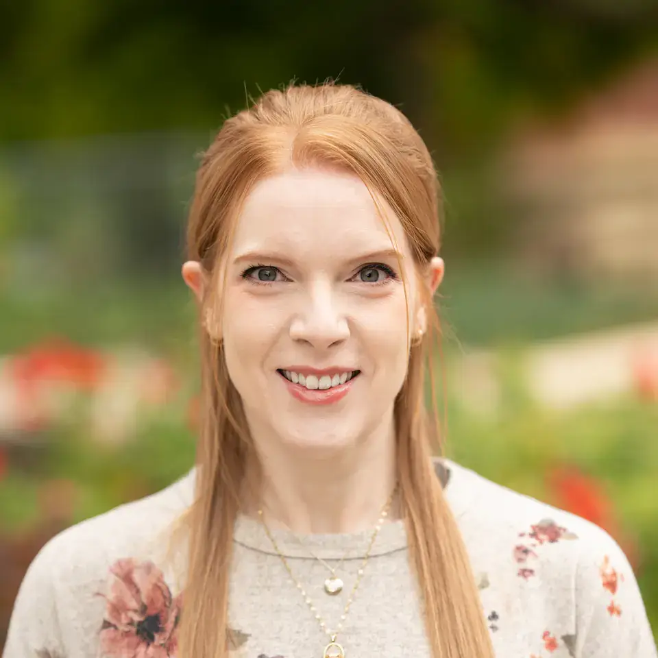 Smiling woman with long red hair in a floral sweater, photographed outdoors against a blurred garden background.