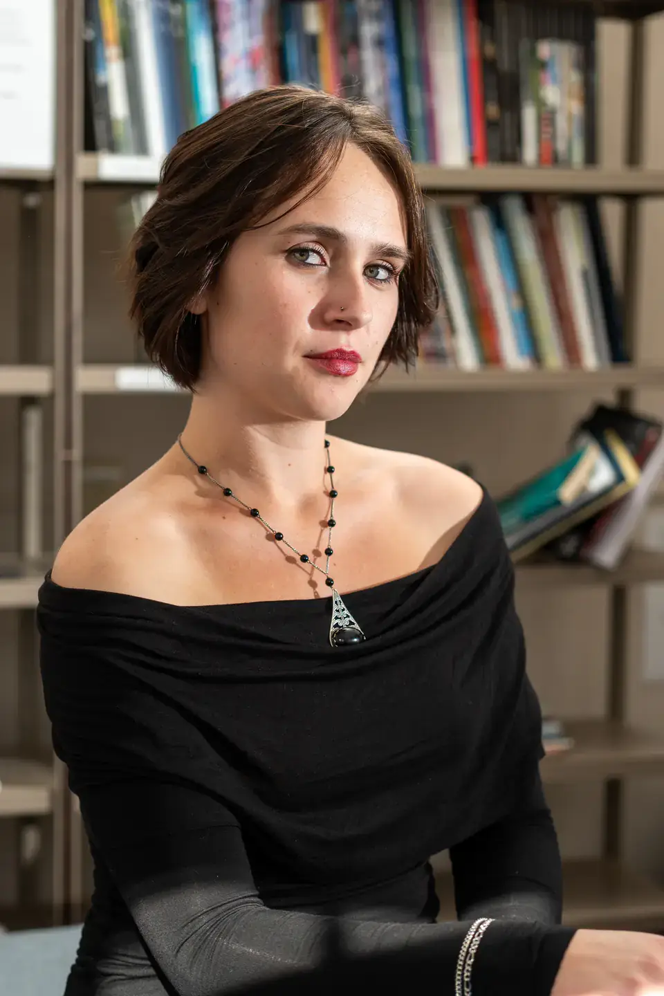Woman with short brown hair in a black off-shoulder dress, seated in front of bookshelves.
