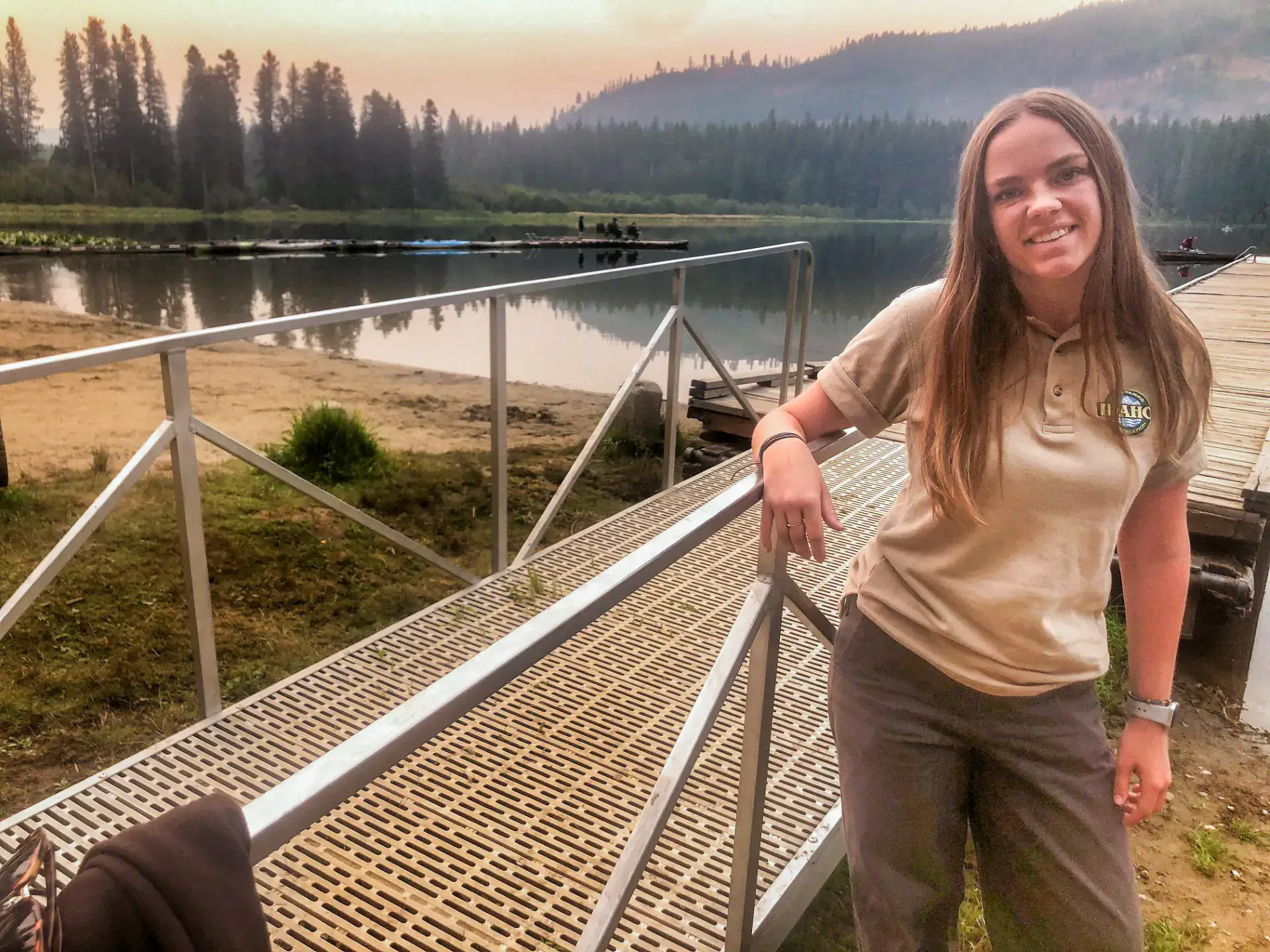 A smiling person stands outdoors by a waterfront dock surrounded by forested hills under an orange, hazy sky.