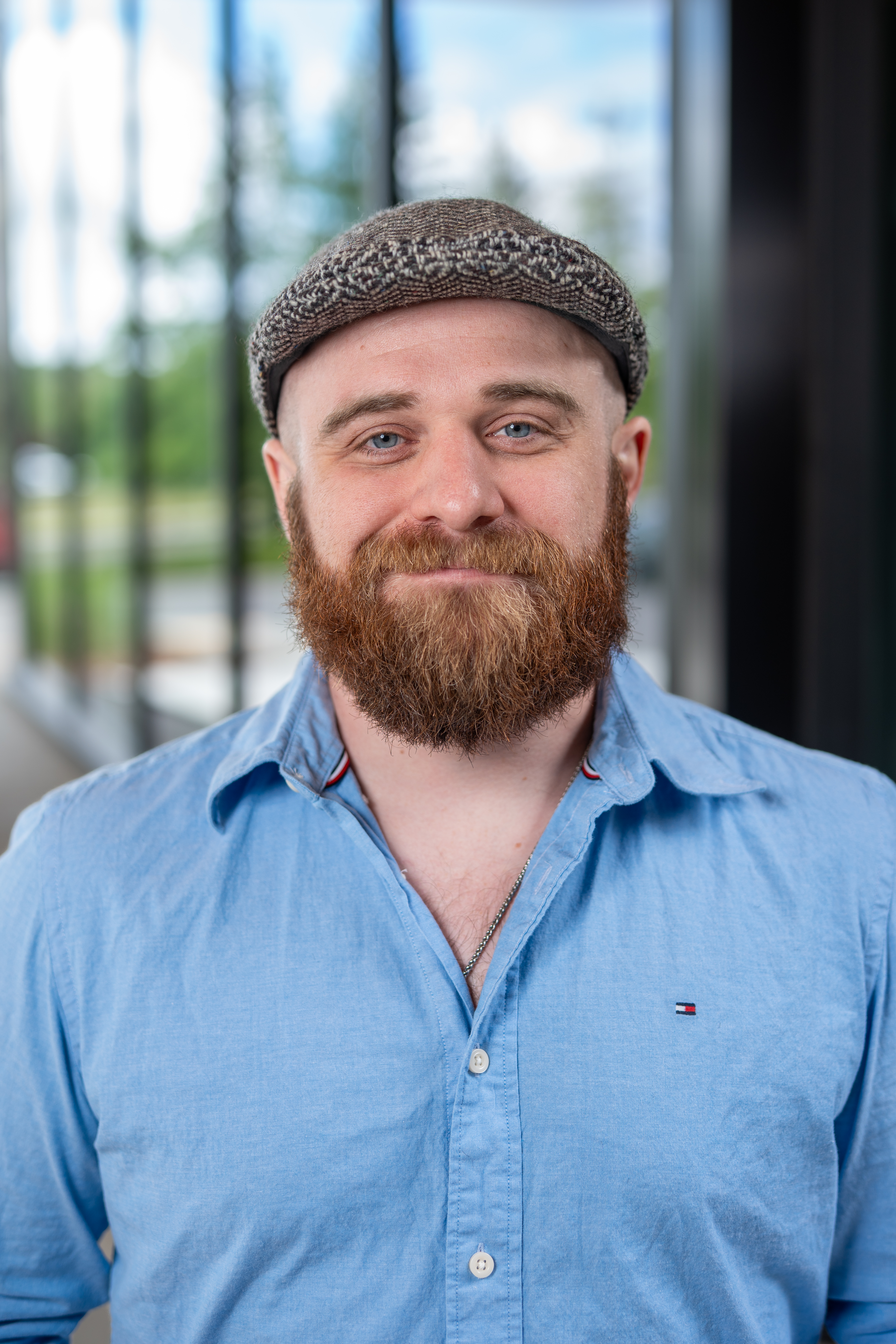A bearded man wearing a light blue shirt and a gray flat cap, with blurred outdoor scenery in the background.