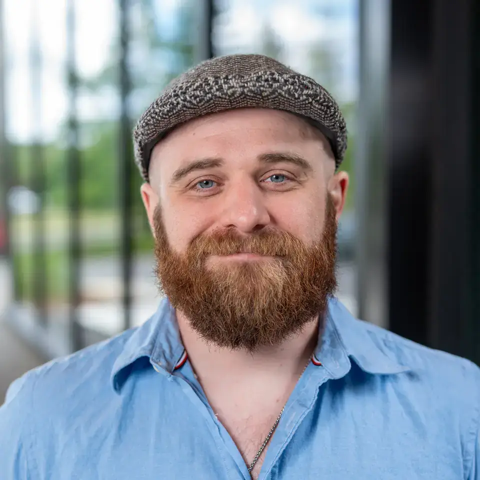A bearded man wearing a light blue shirt and a gray flat cap, with blurred outdoor scenery in the background.