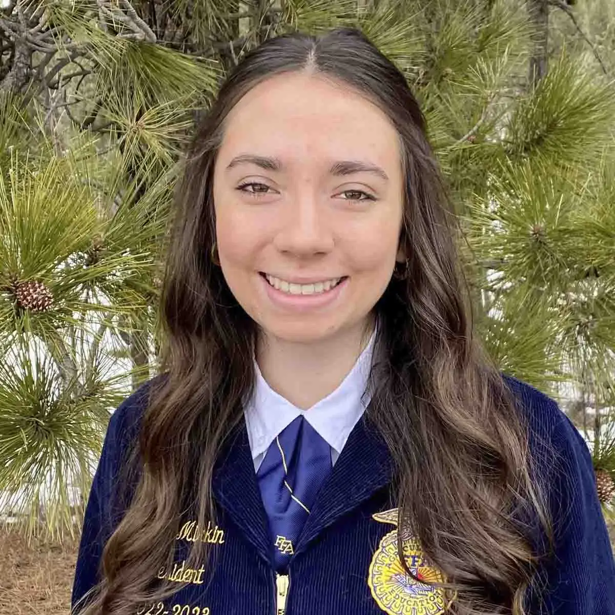 A person in a blue jacket with embroidery representing an organization stands smiling outdoors by a pine tree.