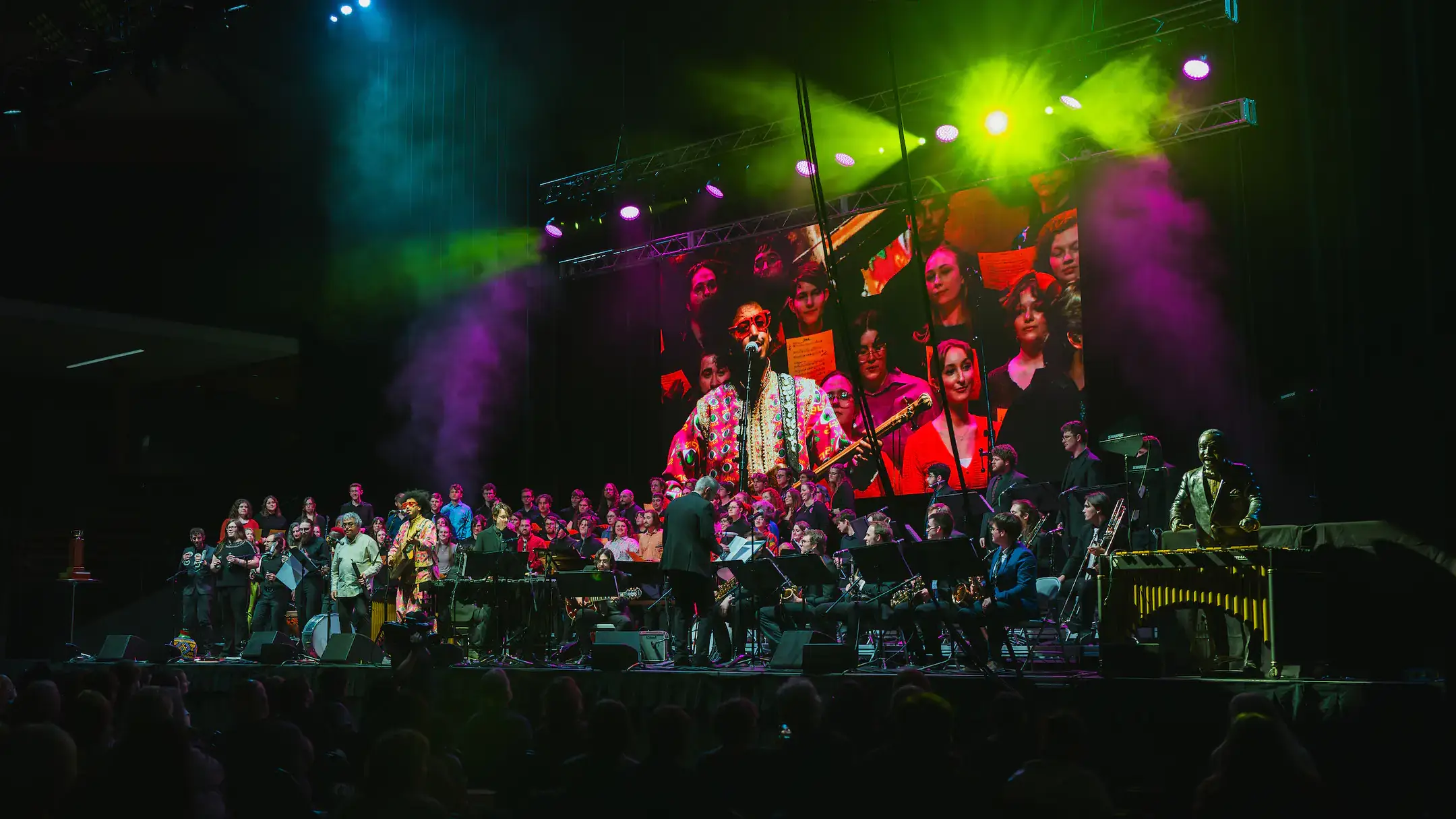 A large group, featuring a vocalist, band and choir,  performs onstage with colorful lighting.
