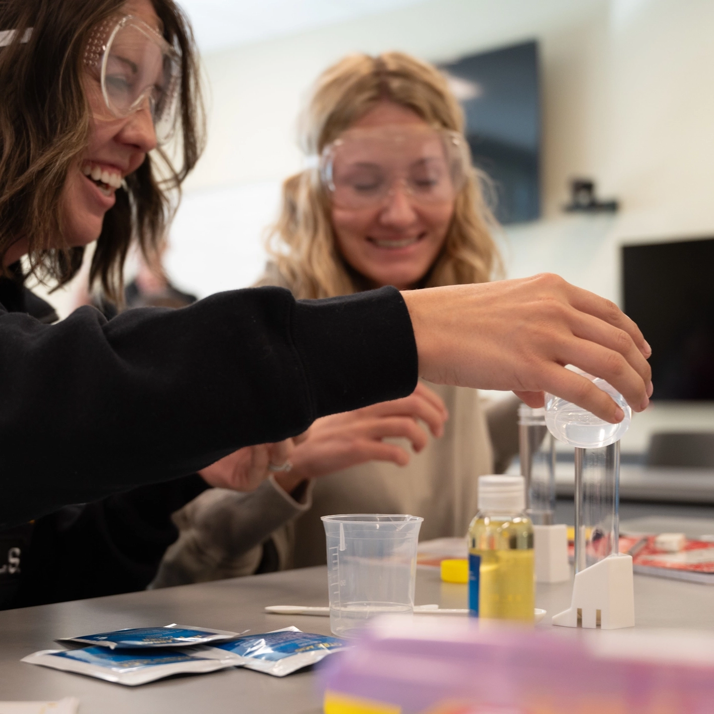 two students wearing safety goggles while one pours liquid into a clear container 