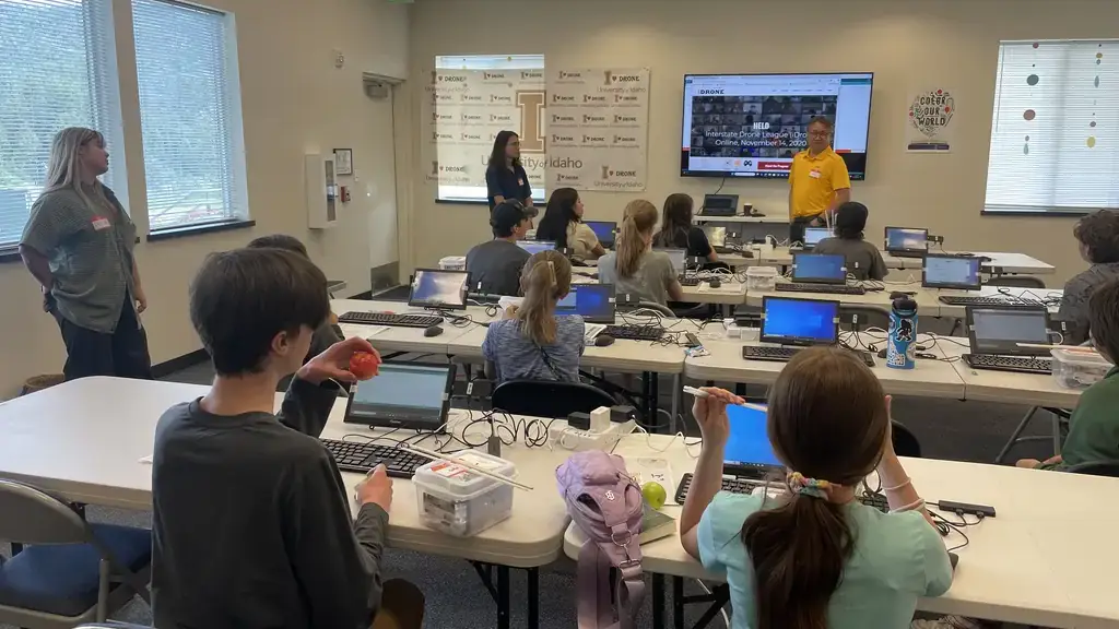 Several youth at rows of tables working with laptop computers listen while Jae Ryu presents a slide presentation by a TV monitor.