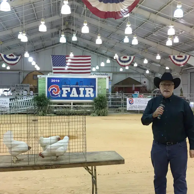 A man wearing a cowboy hat speaks into a microphone in a barn-like setting with American flags and decorative banners. White chickens are in cages resting on a table in the foreground.