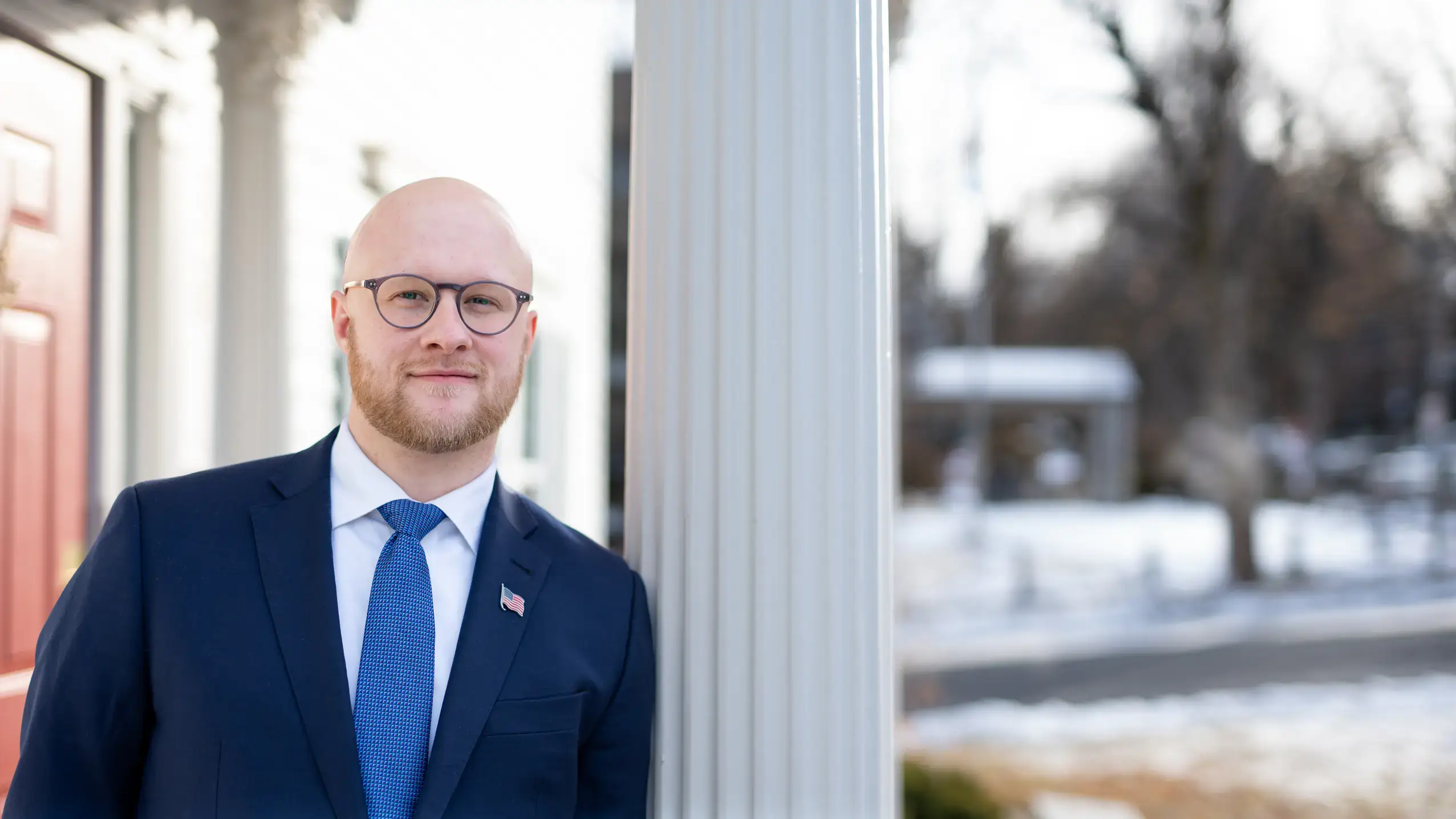 A bearded, bald man with eyeglasses, wearing a suit with an American flag lapel pin stands near a Doric column.