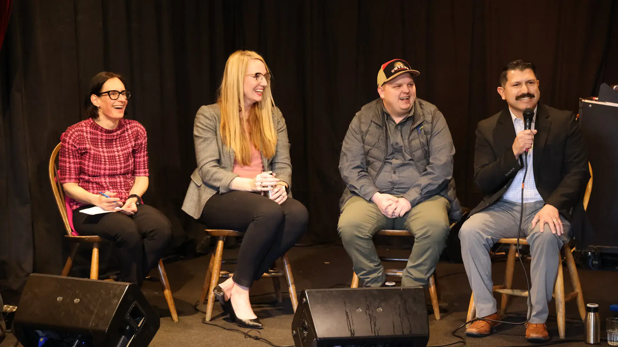 Four people smile while seated on a stage. A man on the far right holds a mic while interacting with the audience.