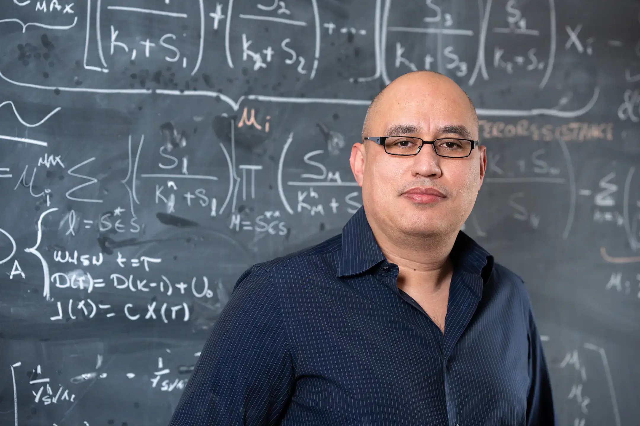Portrait of Assistant Professor of Mathematics Esteban Hernandez-Vargas in front of a chalkboard with math equations models to study the mitigation of viral infections.
