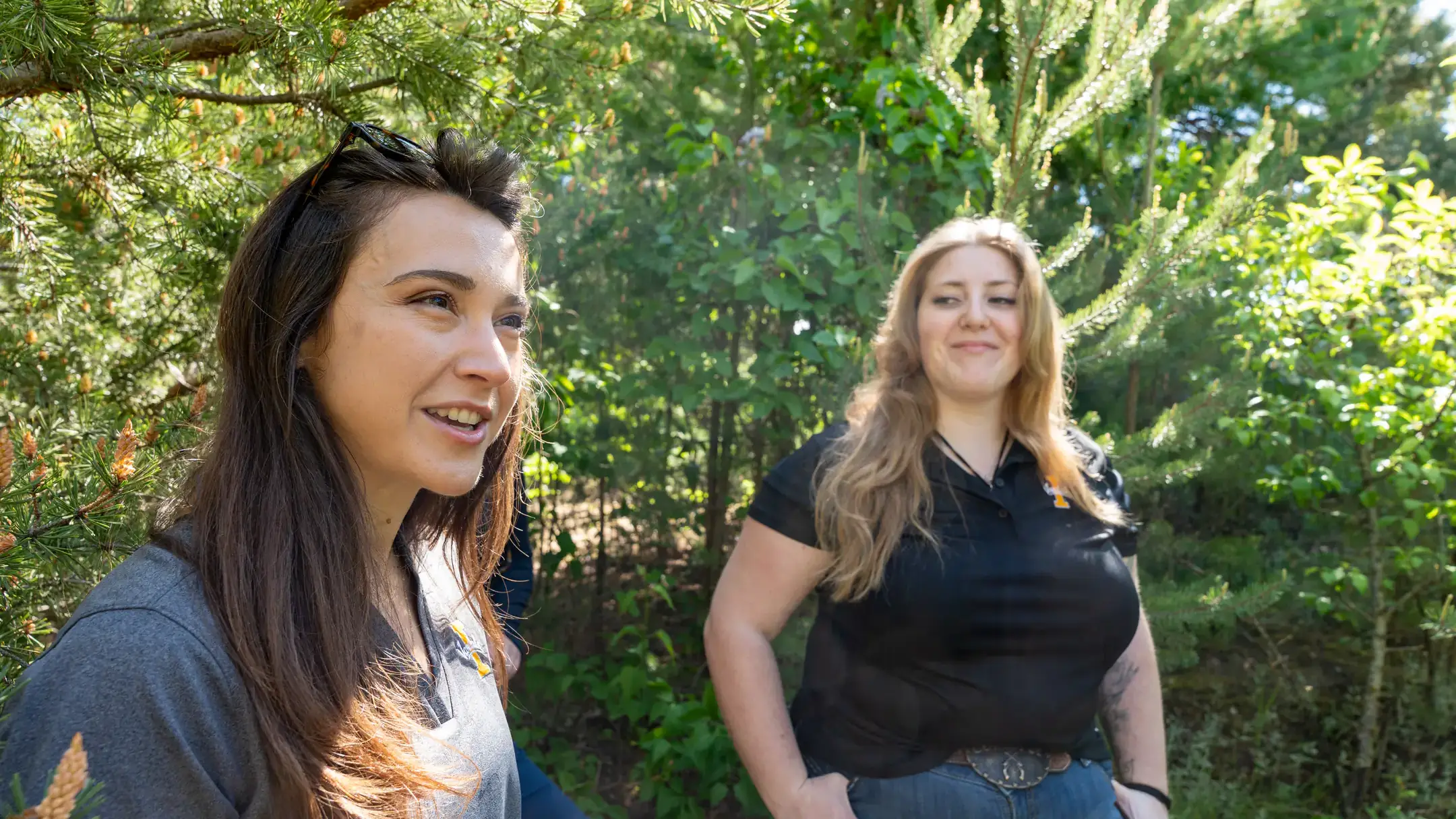 Two people are outdoors in a sunny wooded area, talking and smiling. The background includes trees and foliage.