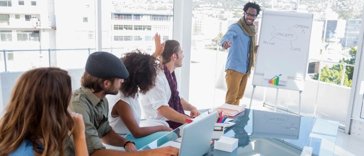 Man standing and pointing at group sitting down at table in office meeting