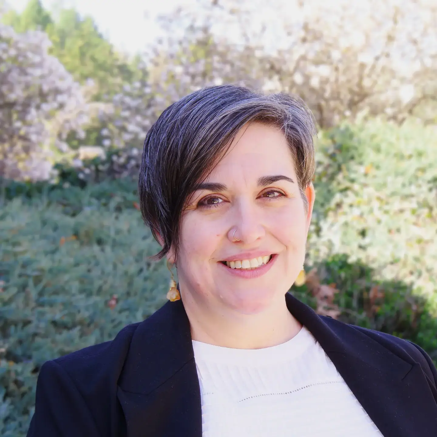 Woman with short hair smiling, in front of a garden background with blooming trees and shrubs.