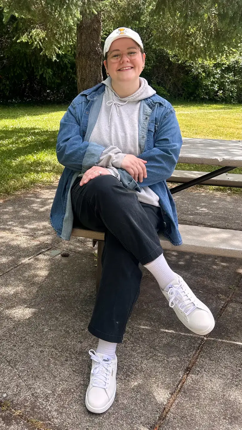  A woman wearing a hat and a denim jacket poses for a photo while sitting on an outdoor picnic bench. 