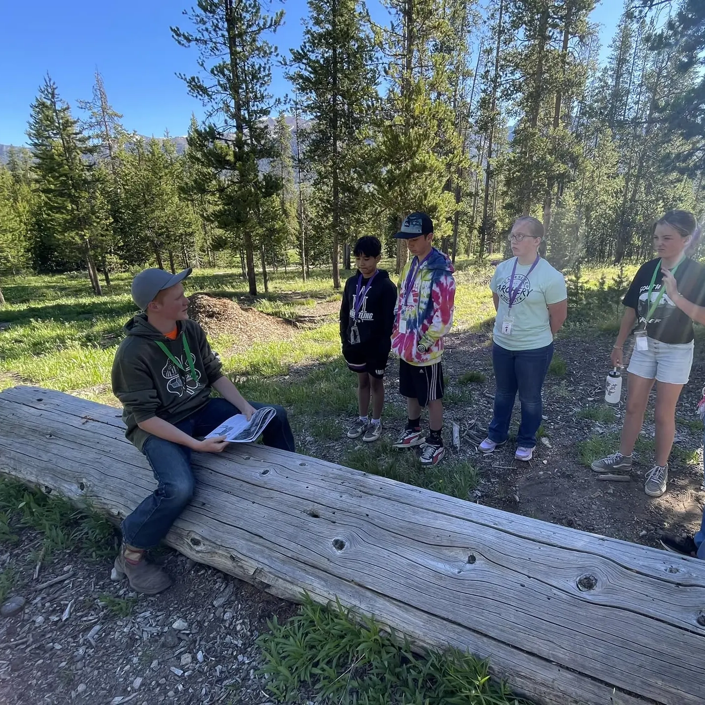 Children stand around instructor sitting on log in the woods.