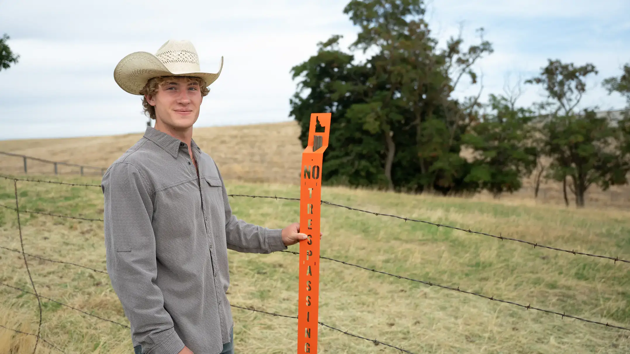 Young man in a cowboy hat holds an orange no trespassing sign by a barbed-wire fence in a rural field.