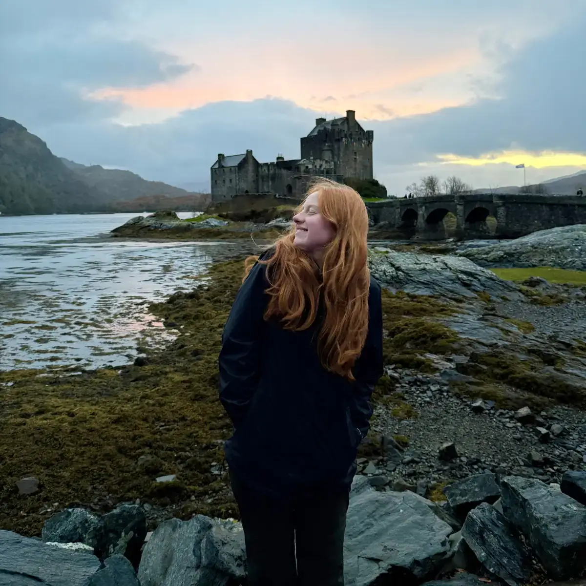 A woman standing in front of a castle that's next to a body of water.