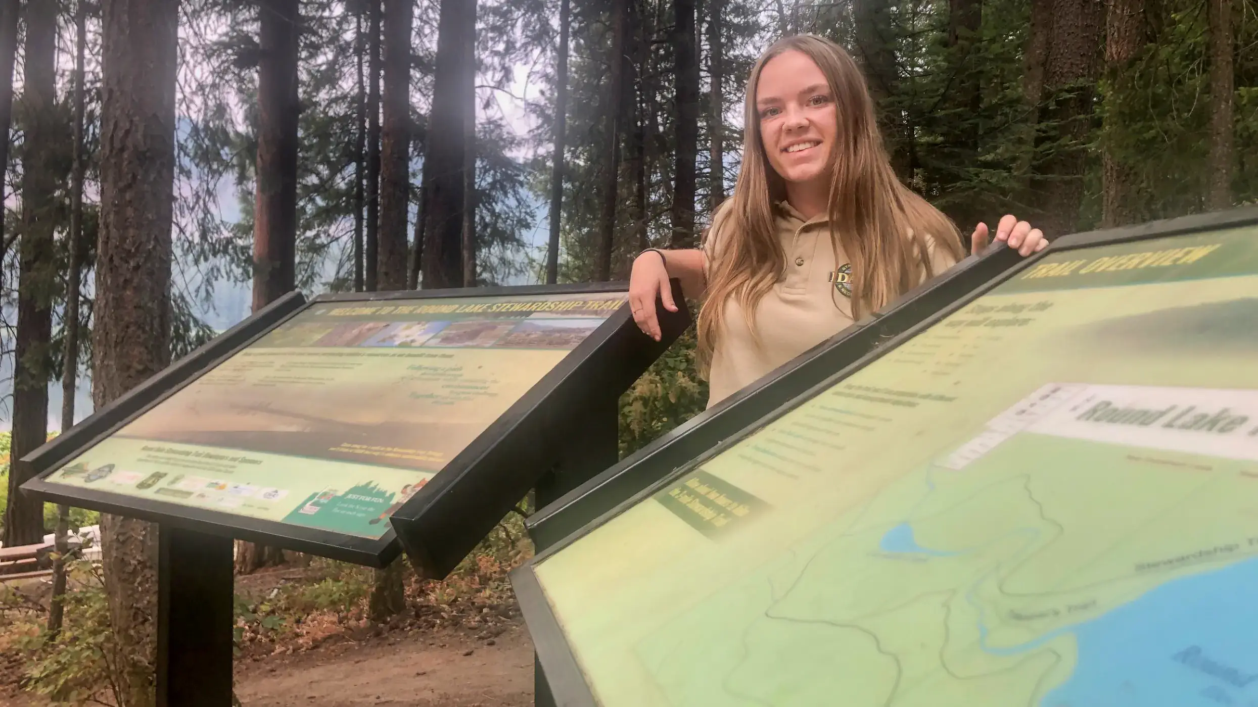 A person stands outdoors near a wooded trail, smiling next to two informational signs about Round Lake.