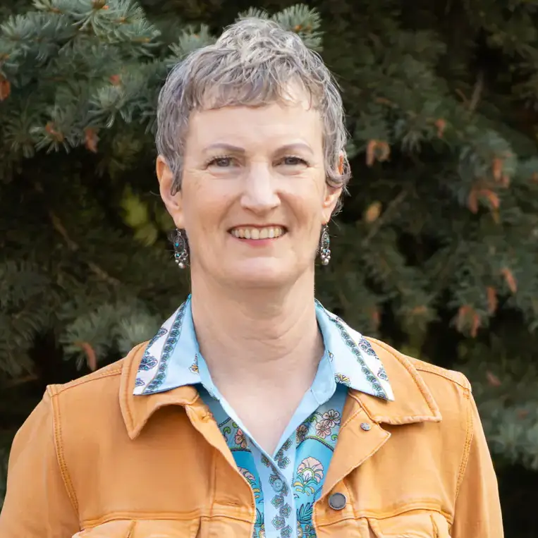 Smiling woman with short gray hair in an orange jacket, standing outdoors in front of evergreen trees.