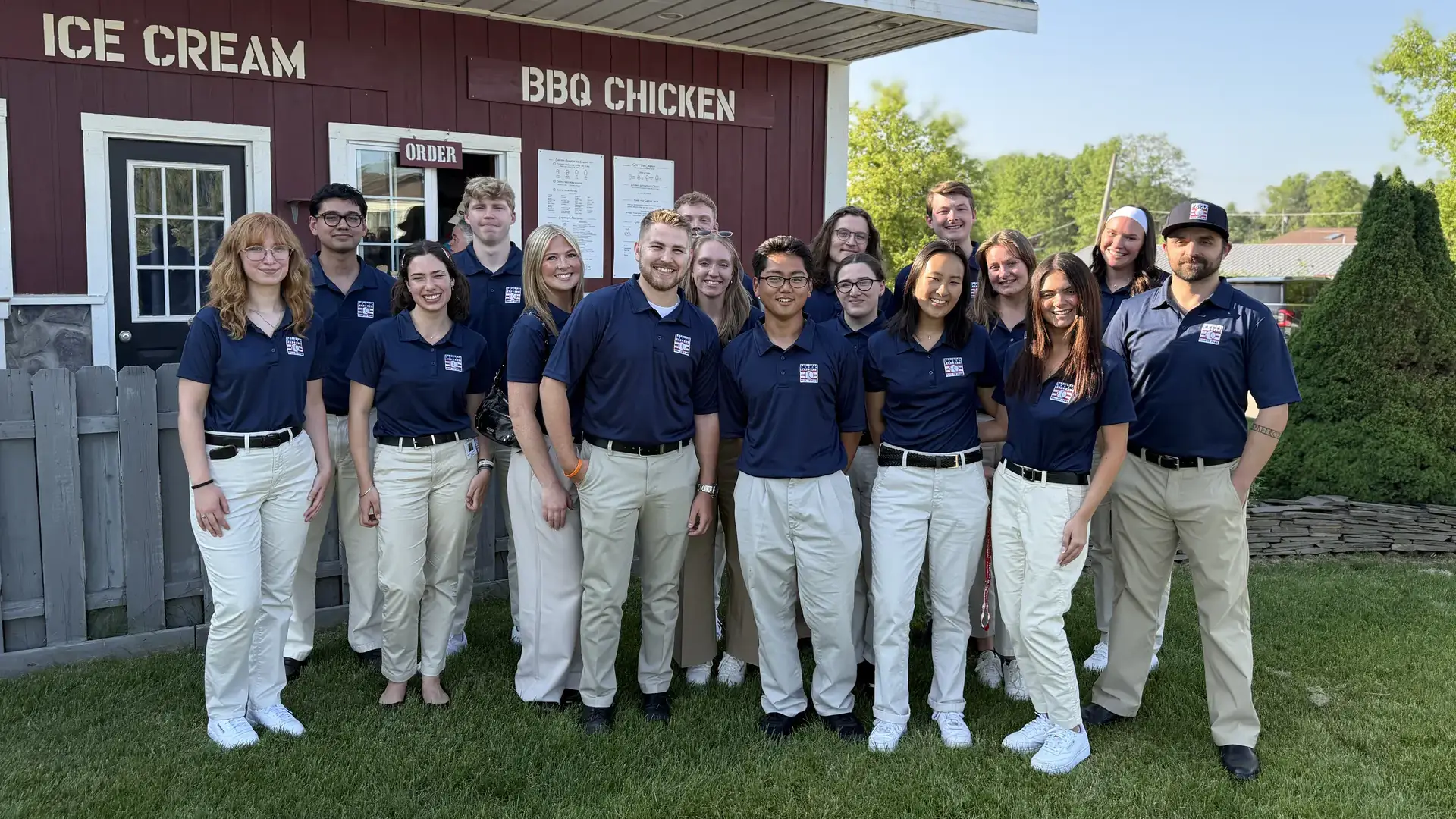 Photo of 17 people wearing blue shirts and white pants standing in front of a concession stand.