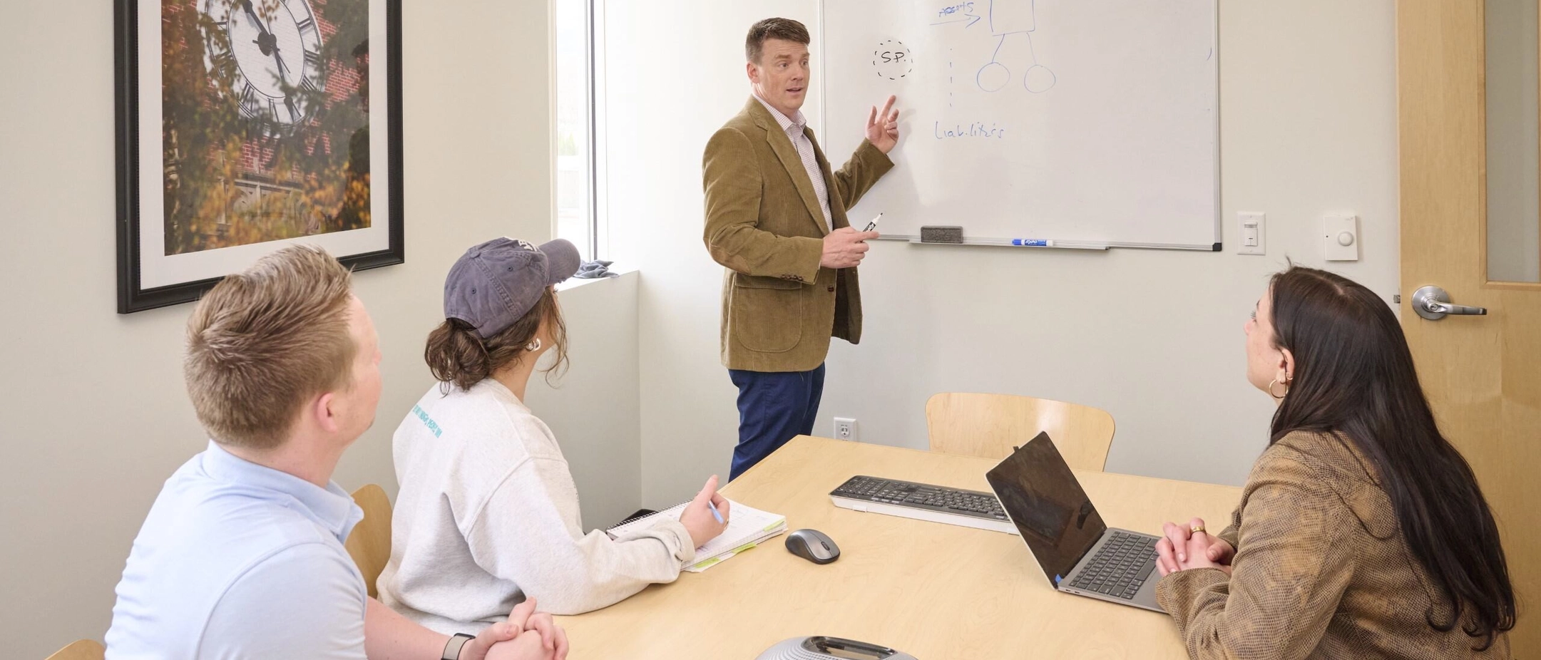 Three people listening and paying attention to a man pointing at a whiteboard