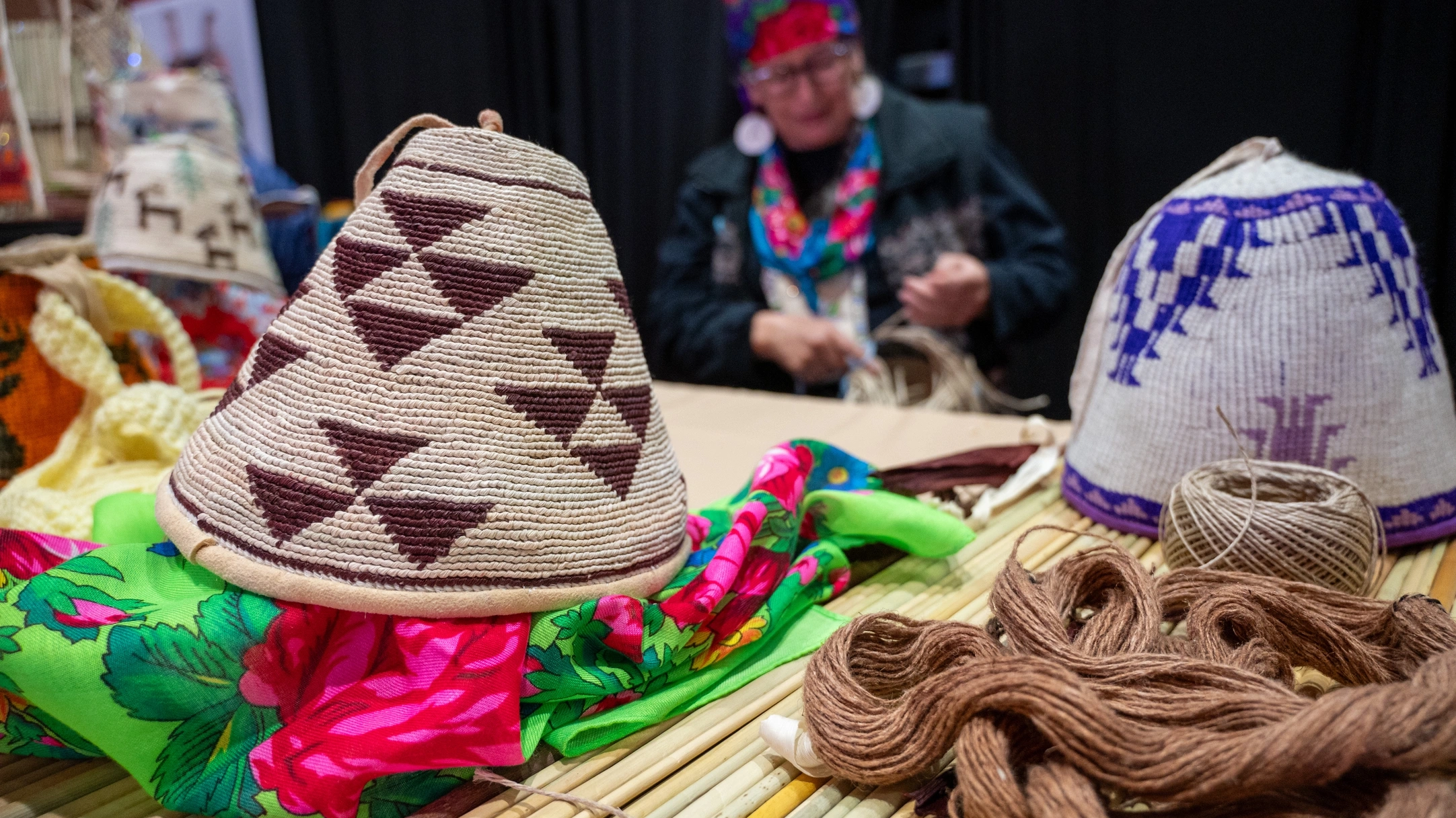 Hand-woven digging bags are displayed on a table while the weaver works in the background.