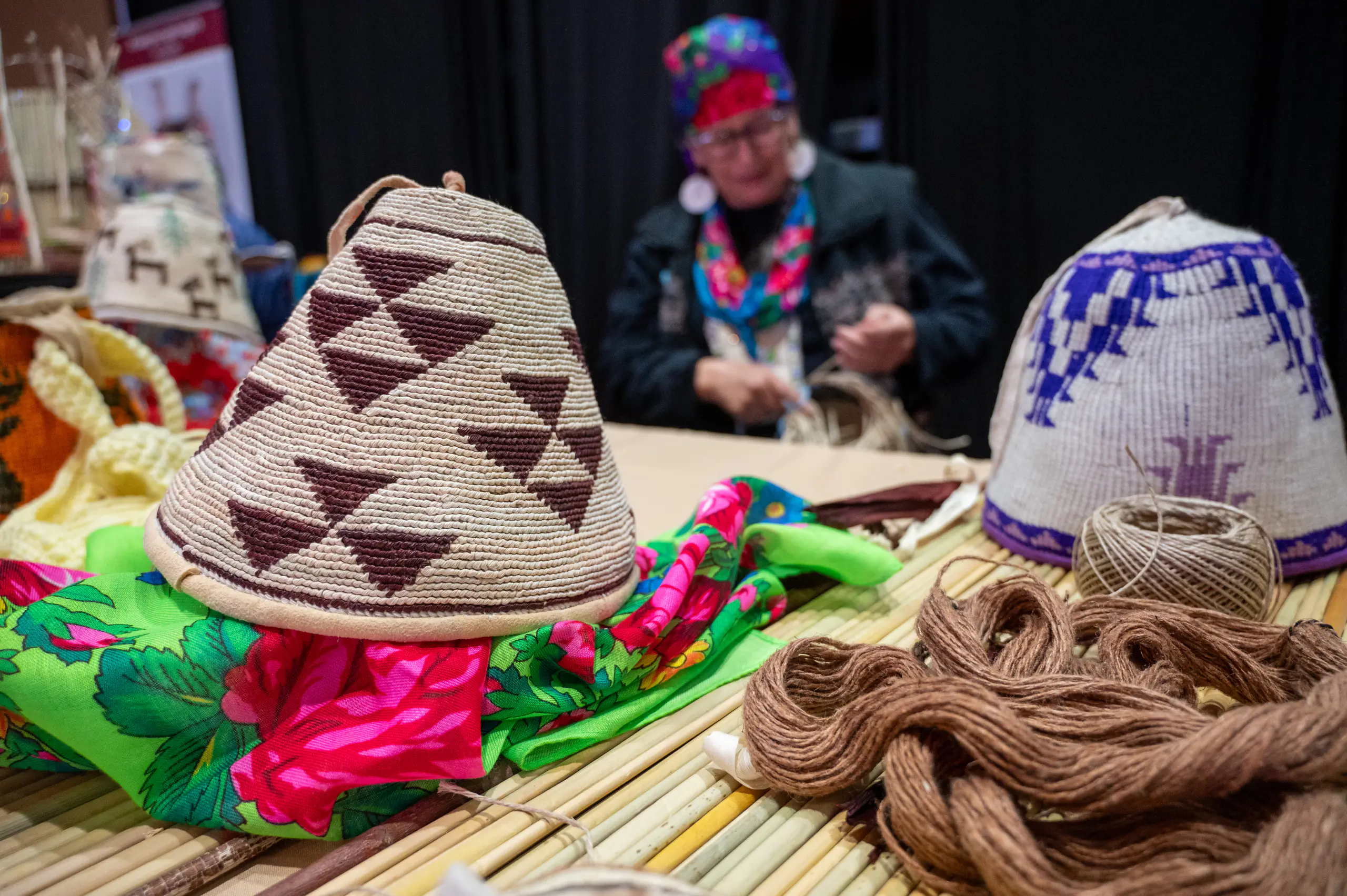 Hand-woven digging bags are displayed on a table while the weaver works in the background.