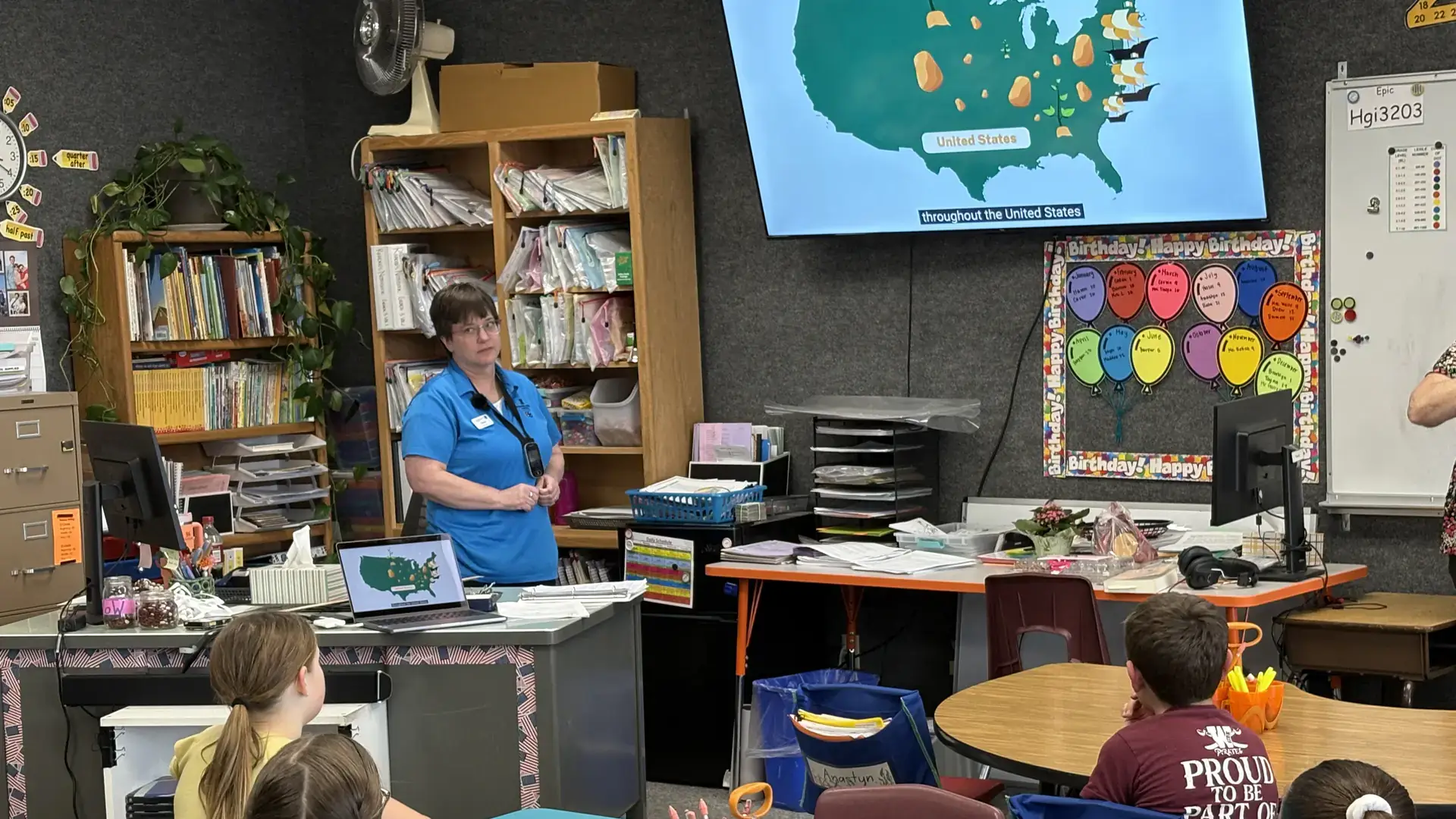 A woman stands by a projection of a world map at the head of a classroom filled with children.