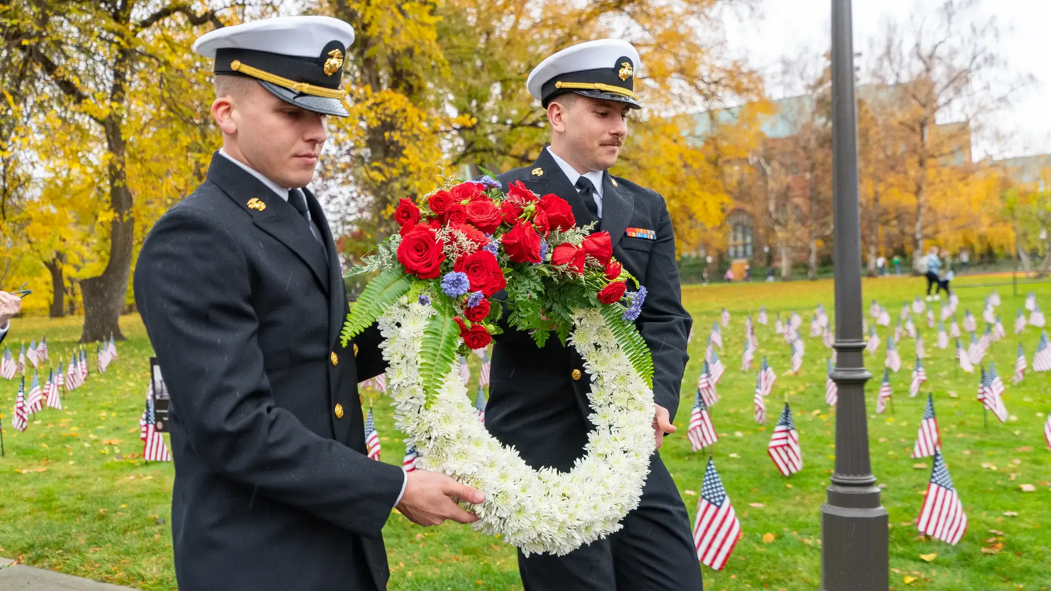 wo people in miliary garb carry a wreath in front of a field filled with small American flags. 