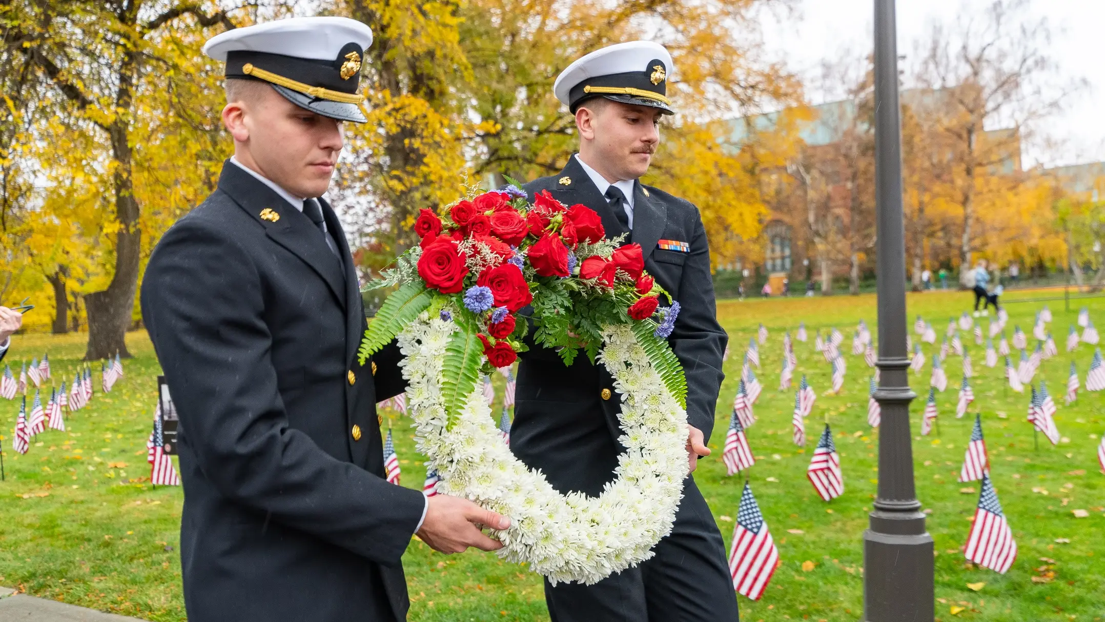 wo people in miliary garb carry a wreath in front of a field filled with small American flags. 