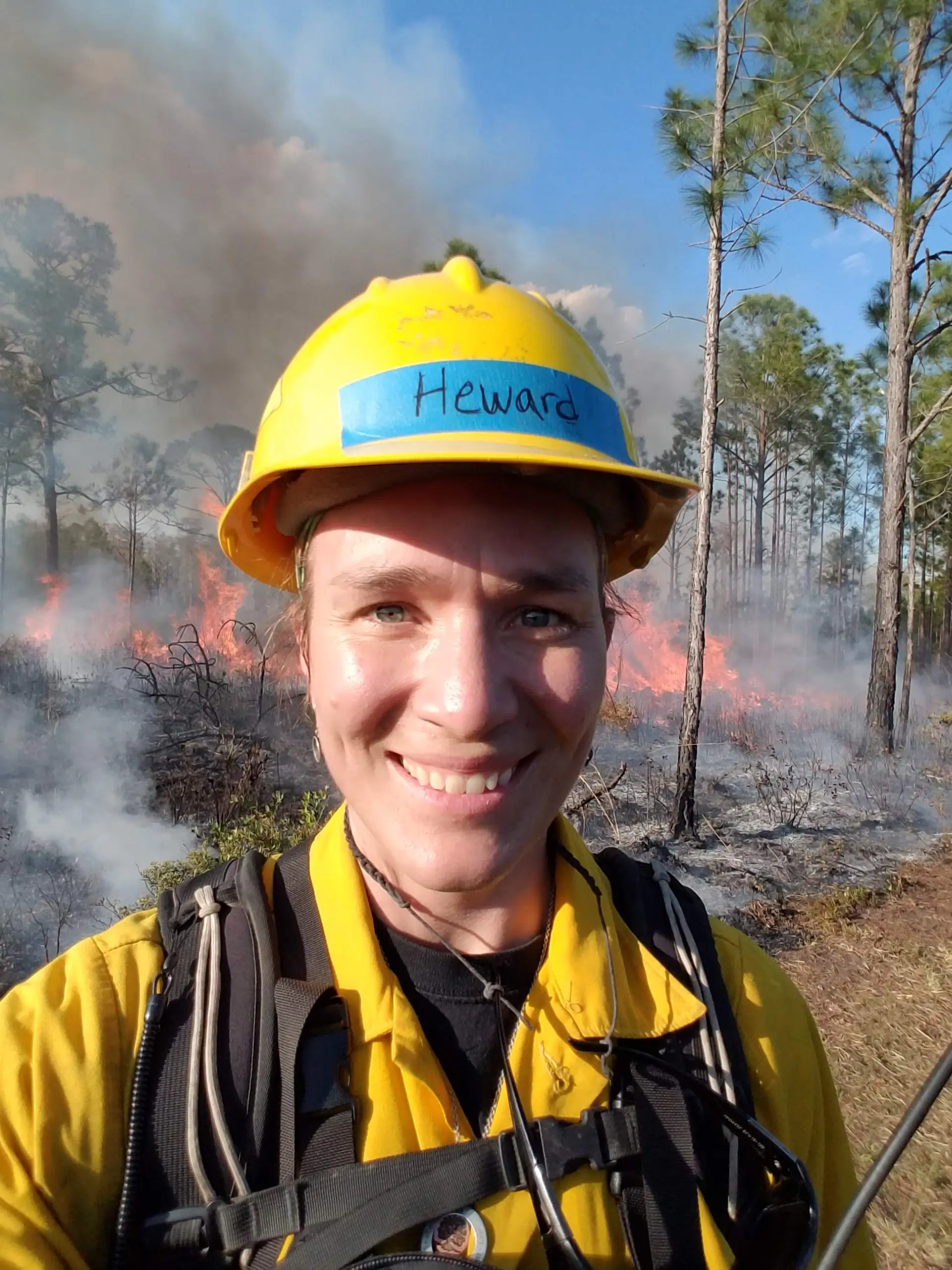 Heather Heward, clad in forest fire fighter gear, smiles with a fire in the background.