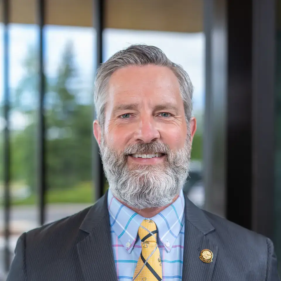 A person wearing a suit, checkered shirt, and a yellow tie with patterned designs, standing outdoors with trees and glass panels visible behind.