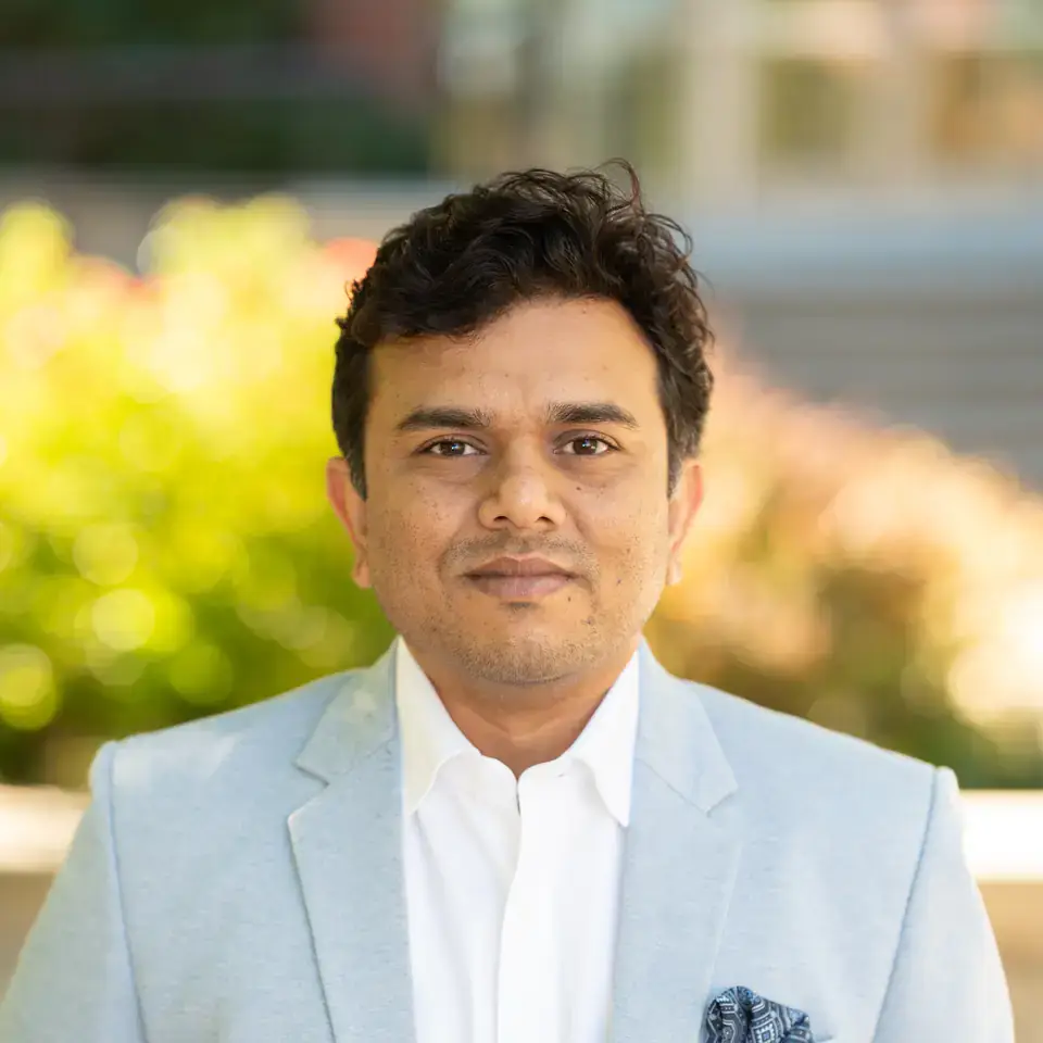 Man in a light blue blazer and white shirt, standing outdoors against a softly blurred background.