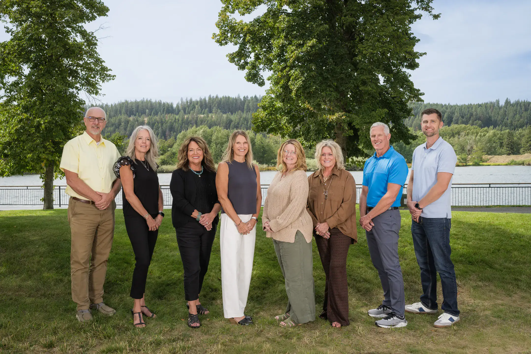 A group of eight individuals standing outdoors in front of trees and a river.
