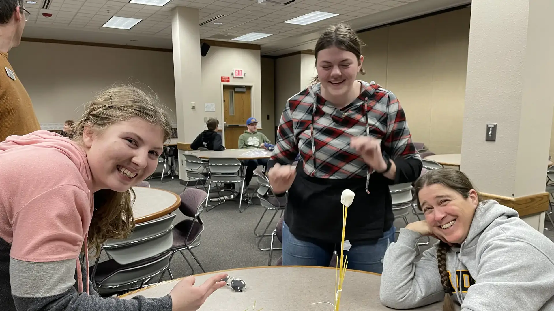 Three women work at a table. One of them, wearing a University of Idaho sweatshirt, holds a sculpture made of spaghetti noodles and marshmallows.