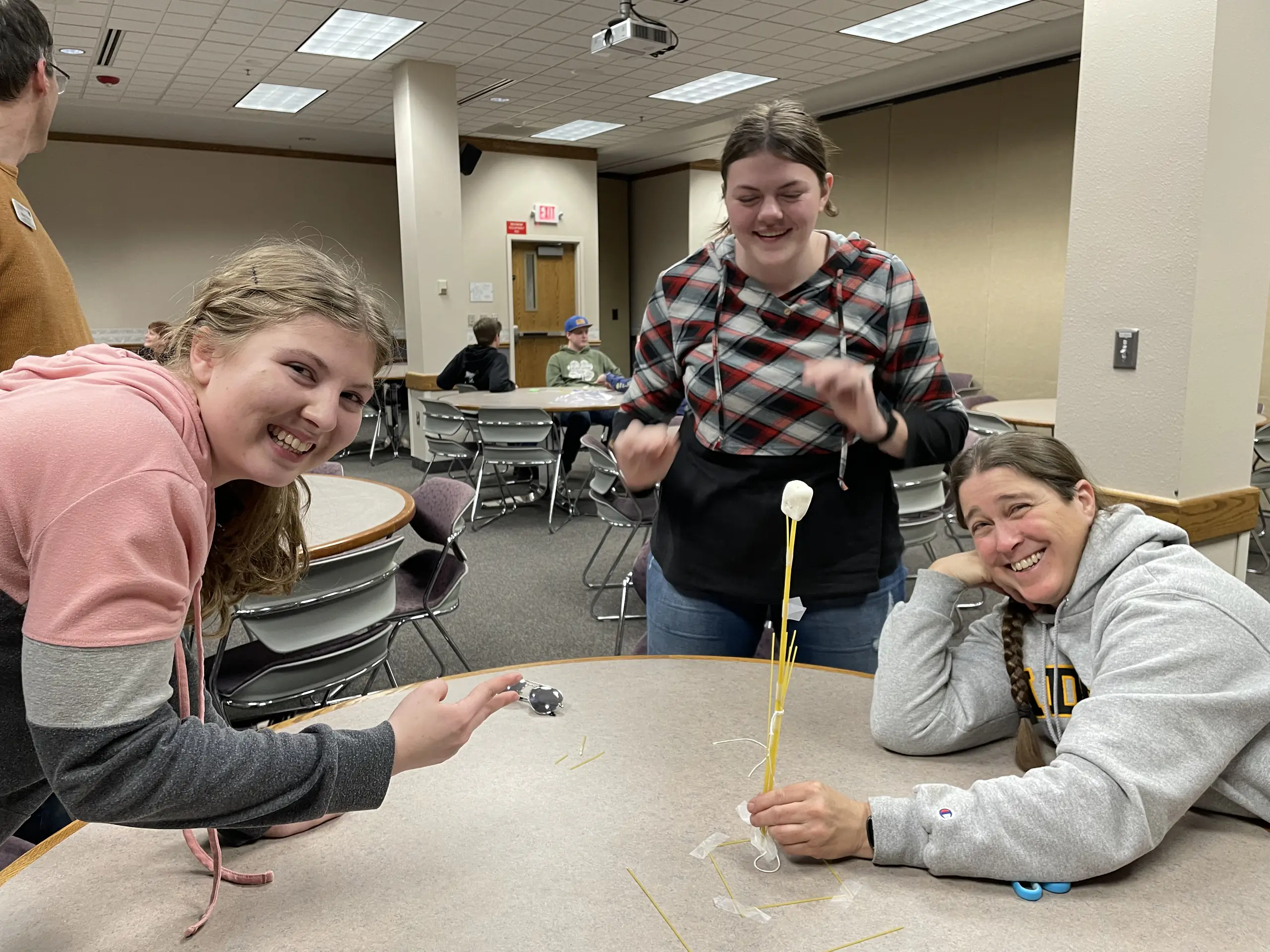 Three women work at a table. One of them, wearing a University of Idaho sweatshirt, holds a sculpture made of spaghetti noodles and marshmallows.