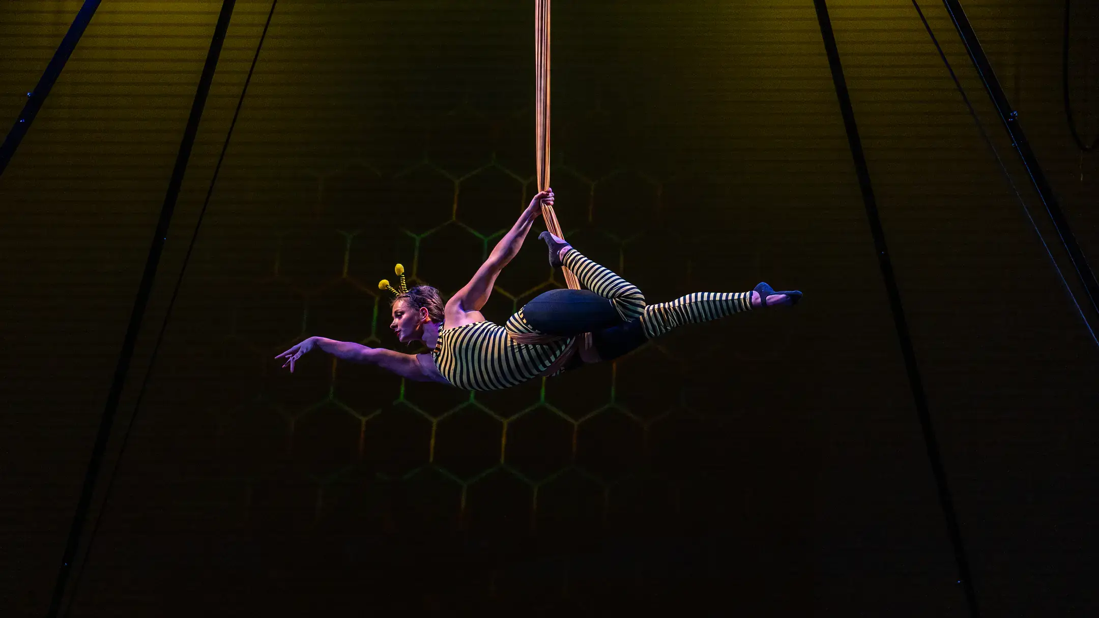 A performer on aerial silks extends horizontally under stage lighting, wearing a striped costume against a dark backdrop.