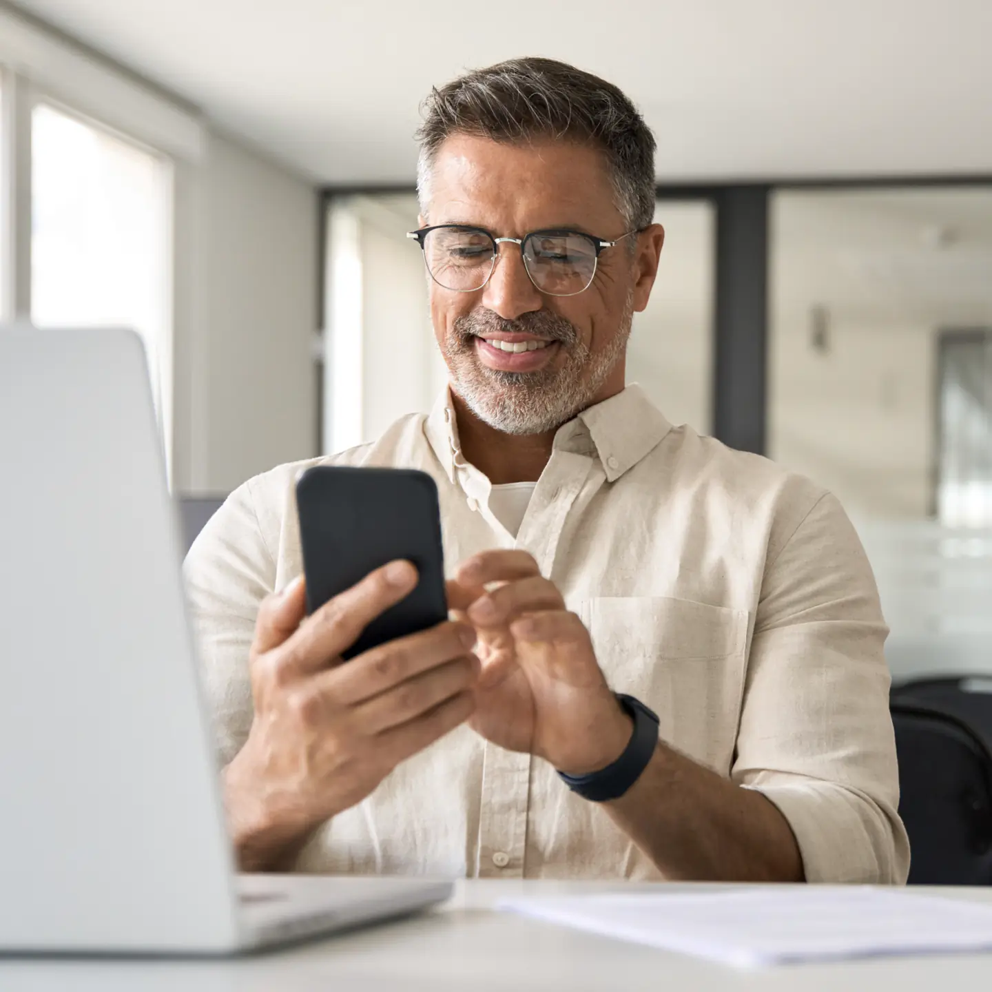 Man smiling while looking at a smartphone, seated next to an open laptop in a modern office setting.