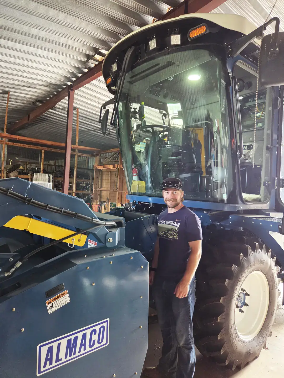 A person standing beside a large agricultural vehicle marked 'Almaco' inside a metal building.