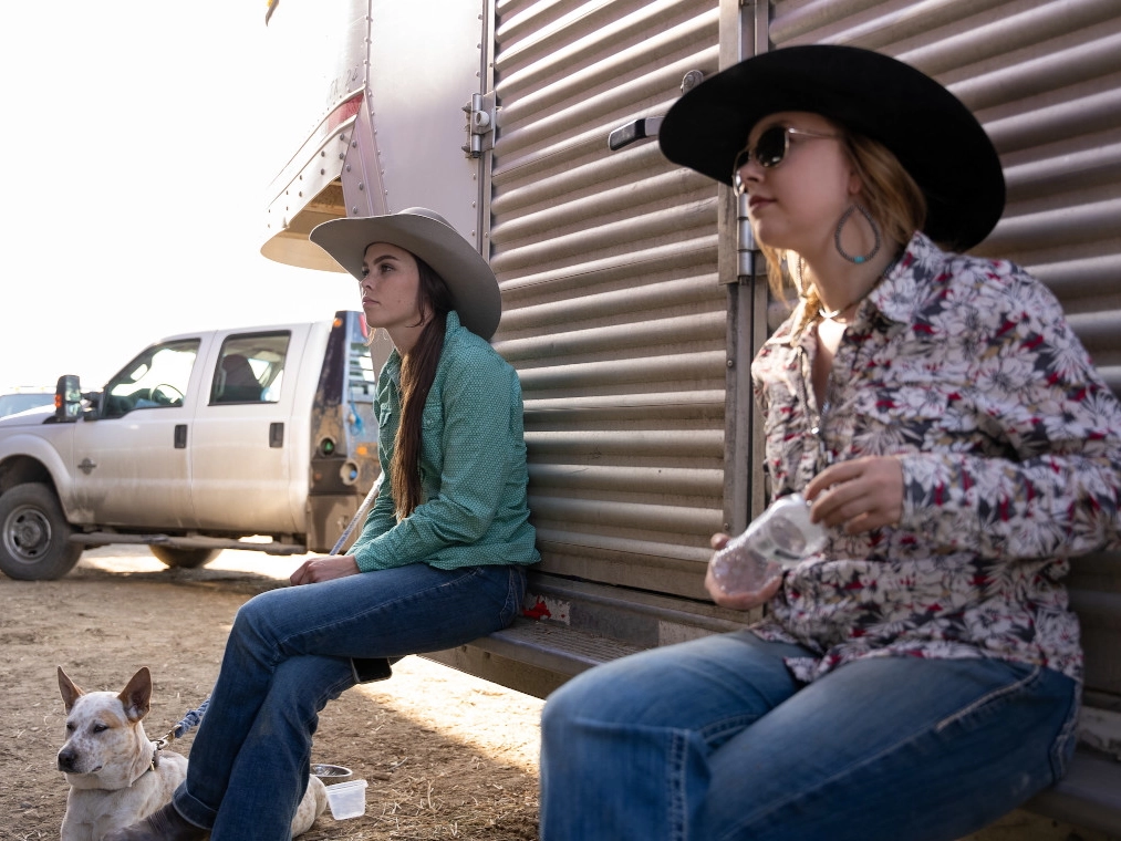 Two women in cowgirl hats sit on the back of a horse trailer