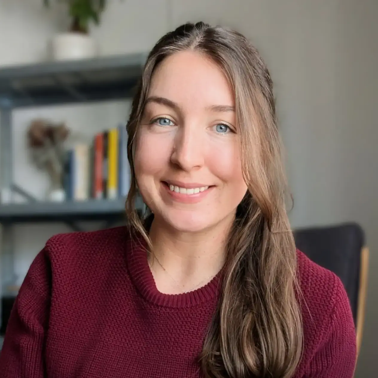 Smiling woman in a burgundy sweater, seated indoors with a blurred bookshelf in the background